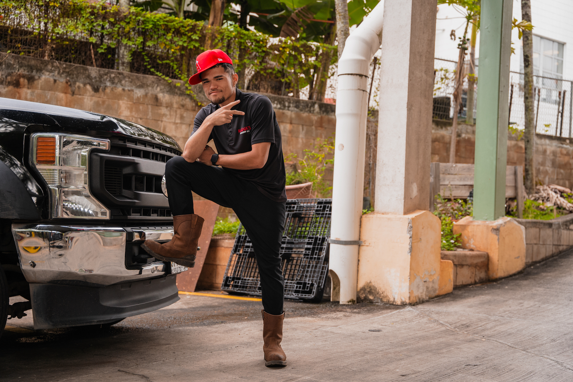 Our employee wearing a red hat and our black uniform with boots on with one of his legs resting up on the bumper of our company dumptruck in Waimalu, Oahu.