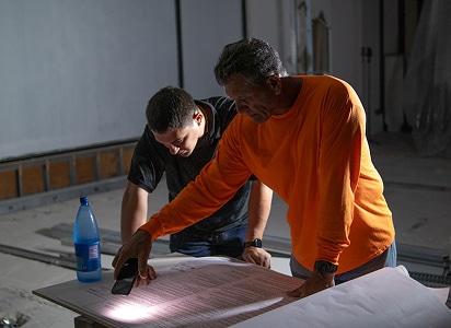 Two men examine a large document on a table, one shining a flashlight on it. The room appears to be under construction or renovation.