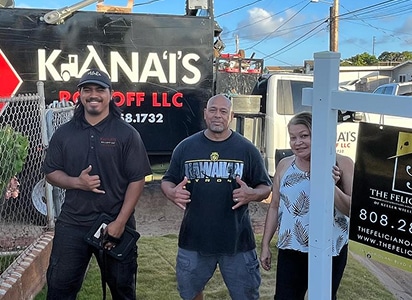 Three people stand outside near a lawn and a fence, with trucks displaying "Kiana'i’s Roll Off LLC" and real estate signage visible in the background.