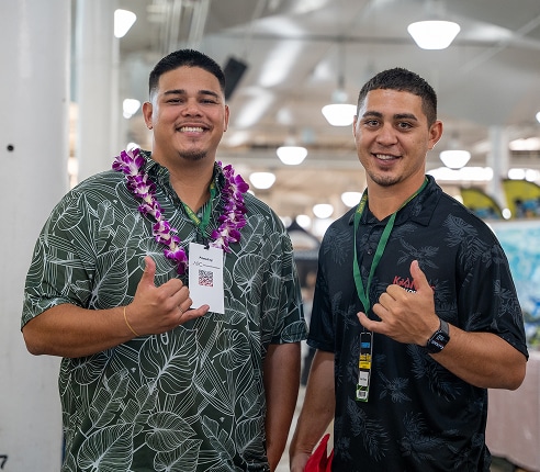 Two men wearing aloha shirts and lanyards stand indoors, smiling and making the shaka hand gesture; one wears a purple lei.