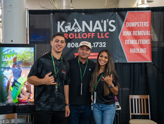 Three people stand smiling in front of a KHANAI'S Rolloff LLC booth with a company banner advertising dumpsters, junk removal, demolitions, and hauling services.