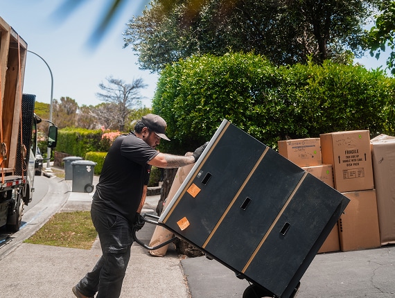 A person uses a hand truck to move a large black cabinet on a residential street, with cardboard boxes and a moving truck nearby.