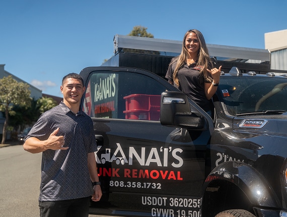 Two people stand by a black truck labeled "Kanai's Junk Removal," both smiling and gesturing shaka signs. One is inside the truck while the other stands beside it.