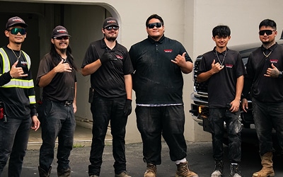 Six men wearing black work uniforms and caps stand outside near a building and a vehicle, posing and making hand gestures.