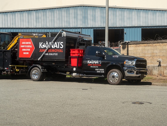 A black Ka'nais Junk Removal truck is parked outside an industrial building, with red bins and equipment on the truck bed.