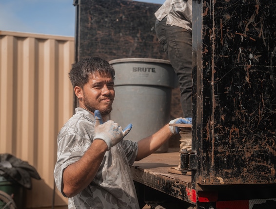 A man wearing gloves stands next to a truck, making a shaka hand gesture, with a trash bin and another person partially visible in the background.