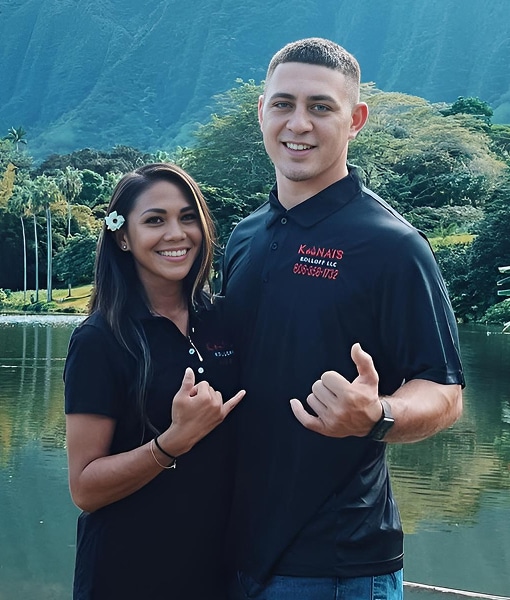 A smiling couple stands side by side by a lake, making shaka hand signs, with lush green mountains and trees in the background.