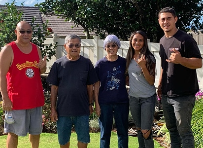 Five people stand together outside in a backyard, smiling at the camera; plants and a fence are visible in the background.
