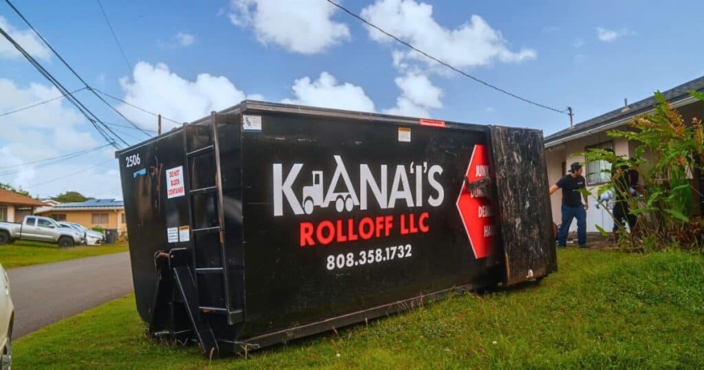 A large black dumpster labeled "Ka'ana's Rolloff LLC" is tipped over on a grassy area near a house, with two people standing nearby.