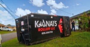 A large black dumpster labeled "Ka'ana's Rolloff LLC" is tipped over on a grassy area near a house, with two people standing nearby.