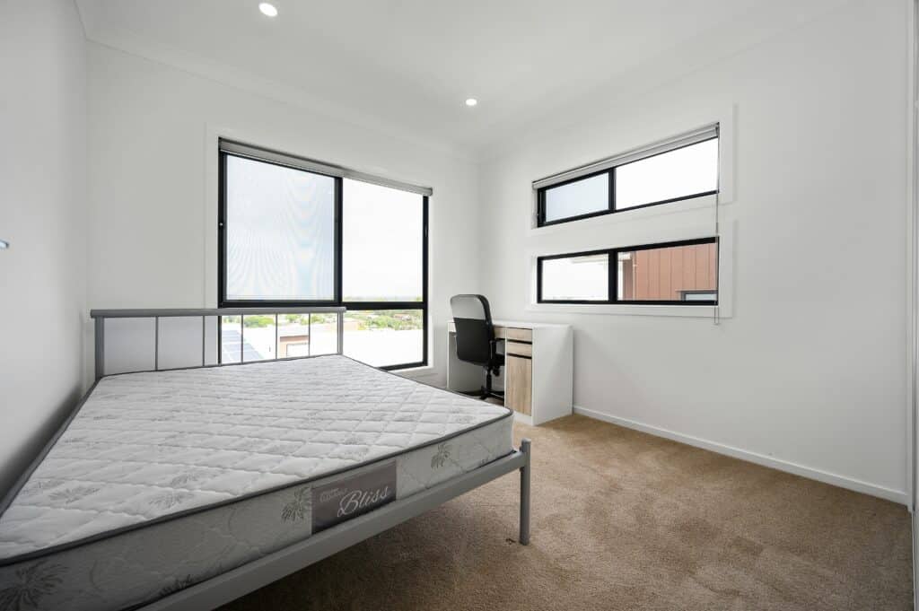 Minimalist bedroom with a metal bed frame, a mattress, a desk with a chair, beige carpet, and large windows providing natural light.